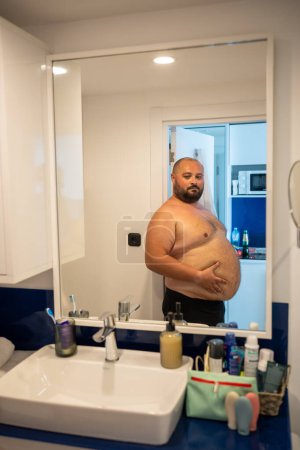Photo for Overweight smiling man with naked torso suffering from extra weight looking at mirror touching abdomen in bathroom at home. Bearded middle aged male inspecting body to change unhealthy life habits. - Royalty Free Image