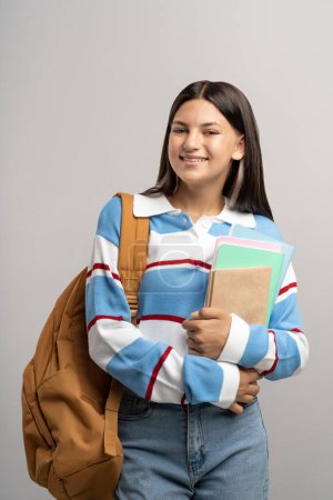 Happy smiling student teen girl with backpack, copy books on gray background. Portrait brunette cute friendly teenager in sweatshirt, jeans. Advertisement banner with copy space, education concept.