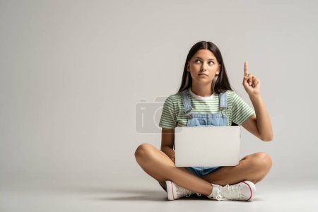 Foto de Chica adolescente inteligente sentado en el suelo con el ordenador portátil que tiene una gran idea, encontrar inspiración, solución al problema. Colegiala positiva señalando con el dedo hacia arriba sobre fondo gris estudio pensando en la idea genio - Imagen libre de derechos