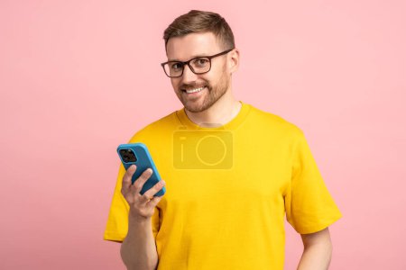 Photo for Pleasant smiling man in glasses with leer sly cunning look holding smartphone after receiving good news, reading desired message, having expected call. Happy man in yellow isolated on pink background - Royalty Free Image