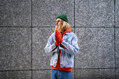 Foto de Retrato rubio delgado chico hipster con el pelo largo, aspecto escandinavo en ropa de mezclilla, guantes naranja sudadera de pie en la calle. LGBT hombre gay transgénero mirando a un lado plegable manos en orar pose - Imagen libre de derechos