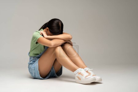 Photo for Frustrated feeling desperate teen girl closing face with hands sitting on gray background. Teenager living through loss of love, heartbreaking. Emotional pain, teenage crisis, depression concept. - Royalty Free Image