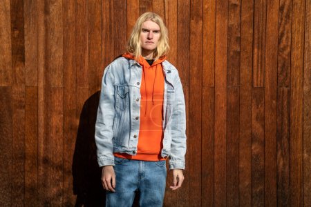 Photo for Apathetic blond long hair man scandinavian androgynous appearance stands near wooden fence looking at camera. LGBT, gay, transgender bisexual guy posing on nature. Individuality personality concept. - Royalty Free Image
