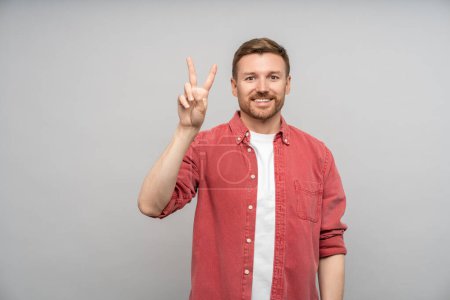 Photo for Smiling positive bearded caucasian guy showing victory sign looking at camera standing on gray background. Portrait middle aged man on poster, banner, placard. Hand gesture, body language concept. - Royalty Free Image