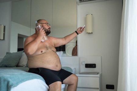 Téléchargez les photos : Homme obèse nu avec bouteille de sueurs d'eau froide beaucoup ajuste le climatiseur avec télécommande en essayant de faire face à des degrés anormaux de chaleur assis dans la chambre d'hôtel. Gros mâle souffre mourir de chaleur. - en image libre de droit