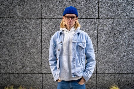 Photo for Portrait blond slim guy with long hair, scandinavian androgynous appearance in glasses looking at camera standing on street with hands in pockets. LGBT man gay transgender in denim clothes blue hat. - Royalty Free Image