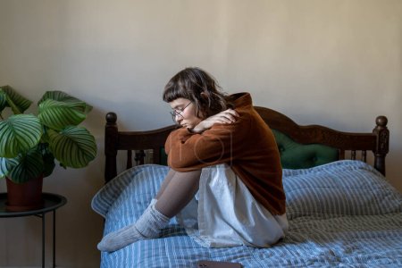 Téléchargez les photos : Apathique adolescent introverti sans émotion assis sur le lit embrassant les genoux seul à la maison. Amabilité, solitude, dépression, absence de motivation, perte d'intérêt pour la vie, concept d'autodestruction - en image libre de droit
