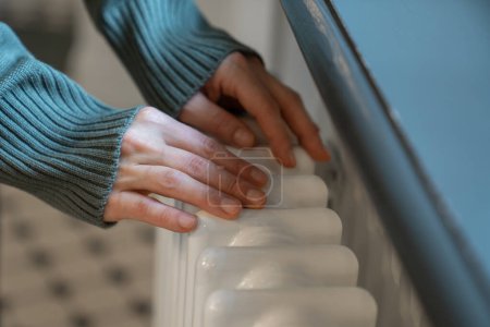 Photo for Cropped photo of woman warming hands near radiator at home, female in warm sweater touching old retro cast iron radiator in winter during heating season, checking heating system working capacity - Royalty Free Image
