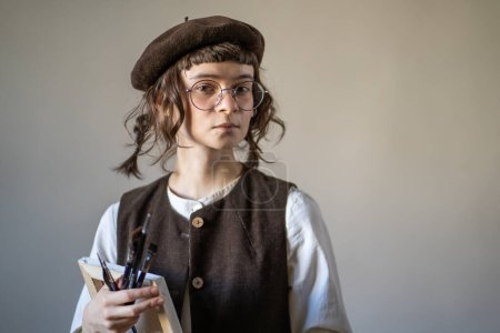 Photo for Serious illustrator in vintage clothes holding canvas, paint brushes, looking at camera. Young artist preparing for work, drawing, creating sketches. Profession of drawing tutor, illustrator, painter - Royalty Free Image