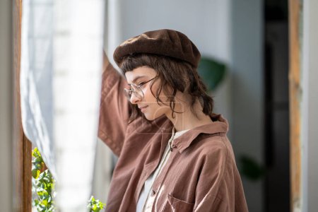 Photo for Pensive melancholic teenager in vintage clothes, beret and glasses standing at window ang looking attentively. Teen girl with concentrated glance looks like dreamer, bookworm, brainy diligent student - Royalty Free Image