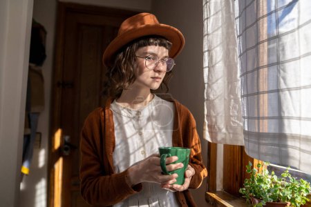 Téléchargez les photos : Adolescente sérieuse dans le chapeau tenant une tasse de thé debout à la fenêtre et regardant la caméra. Adolescent dans de grands verres avec un regard concentré ressemble étrangement fille rétro. Ombre solaire tombant sur le visage - en image libre de droit