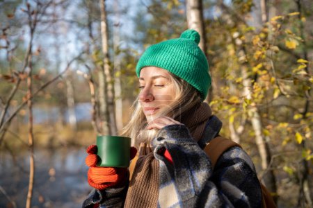 Photo for Smiling of pleasure tourist woman standing in wood with cup of hot tea, relaxing after hiking in sun rays with eyes closed, breathing fresh air, listening to birds singing, enjoying pastime in nature - Royalty Free Image