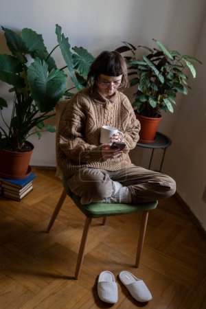 Photo for Introverted teenager spending time alone at home, reading news in internet, scrolling social networks, messaging in smartphone. Teen girl sitting in cozy room with houseplants drinking cup of tea - Royalty Free Image