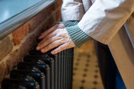 Photo for Cropped photo of woman warming hands near radiator at home, female touching old retro cast iron radiator in winter during heating season, checking heating system working capacity - Royalty Free Image
