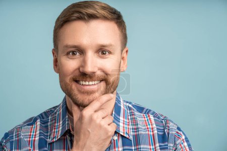 Photo for Portrait studio shot caucasian bearded man looking at camera smiling touching chin on blue background with copy space in right. Positive blond male feeling joy, optimism. Relaxed guy enjoying life. - Royalty Free Image