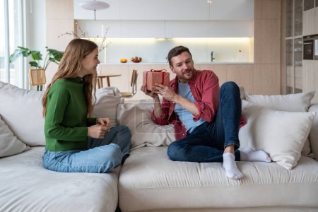 Photo for Joyful woman giving surprise gift present box to man congratulating sitting on couch at home. Happy couple celebrating birthday, anniversary, family holiday together. Loving spouses enjoying marriage. - Royalty Free Image