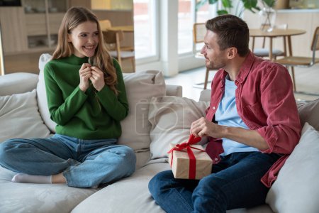 Photo for Joyful wife looks at husband unpacking present gift box smiling expecting reaction sit on couch at home. Woman congratulating man. Happy couple celebrating birthday anniversary family holiday together - Royalty Free Image