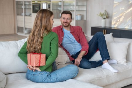 Photo for Interested man look at woman have surprise hiding gift box behind back sit on couch at home. Happy couple celebrating birthday, anniversary, family holiday together. Loving spouses enjoying marriage. - Royalty Free Image