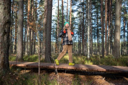 Foto de Mujer de mediana edad turista caminando descansando en sendero ecológico en el bosque de pinos escandinavo a lo largo de camino de madera. Mujer relajada disfrutando del pasatiempo en unidad con la naturaleza. Viajes turismo wanderlust concepto de vacaciones. - Imagen libre de derechos
