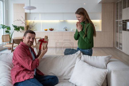 Photo for Interested happy man shaking gift present to understand what inside woman sitting on couch near with folded palms. Pleased wife congratulating surprised husband. Celebrate birthday anniversary holiday - Royalty Free Image