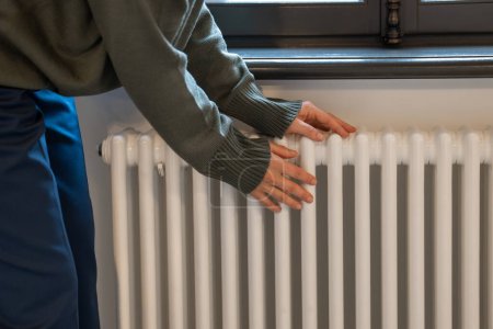 Photo for Woman warming hands near radiator at home after walking in cold winter weather, female touching barely warm battery during heating season, person near window checking heating system - Royalty Free Image