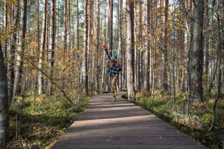 Foto de Mujer alegre caminando saltando levantando la mano regocijándose en el bosque de otoño. Las hembras positivas disfrutan del pasatiempo en el sendero ecológico en el santuario de vida silvestre del parque natural escandinavo. Viajes turismo wanderlust. - Imagen libre de derechos