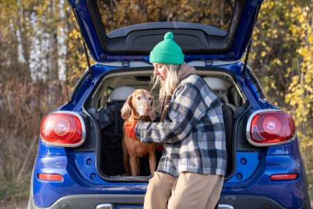 Photo for Loving middle-aged pet owner travelling with purebred hound dog magyar vizsla in car. Female sitting in automobile boot during stop for meal, hugging beloved adorable puppy. Spending pastime with pets - Royalty Free Image