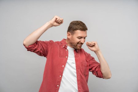 Photo for Happy man rejoices raising hands fists up. Middle aged guy in red shirt smiling laughing happily dancing, rejoicing, celebrating with hands up. Success, luck, joy. Isolated on gray, studio portrait. - Royalty Free Image