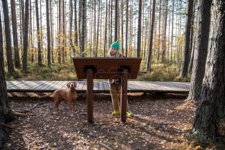 Foto de Turista mujer de mediana edad caminando por sendero ecológico leyendo información sobre el stand en el bosque natural escandinavo. Propietario de mascotas que viaja con un amigo perro disfrutando del bosque con pinos. Turismo de viajes - Imagen libre de derechos