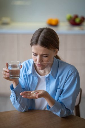 Photo for Woman suffering depression taking antidepressants neuroleptics drinking water sitting on kitchen at home. Sad female treating with pills, tablets. Mental illness, personality disorder, endogenous sick - Royalty Free Image