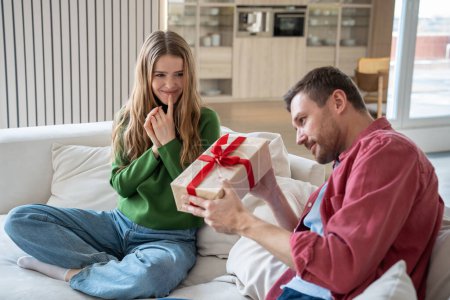 Photo for Pleasantly smiling merry European woman looking with excitement as intrigued interested man trying to unwrap giftbox with red ribbon. Unexpected surprise for birthday, anniversary, New Year - Royalty Free Image