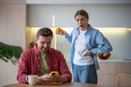 Photo for Jealous controlling wife reads husband messages in cellphone. Infidelity mistrust marital discord concept. Amazed shocked woman standing behind back peeping in mans smartphone on kitchen at home. - Royalty Free Image