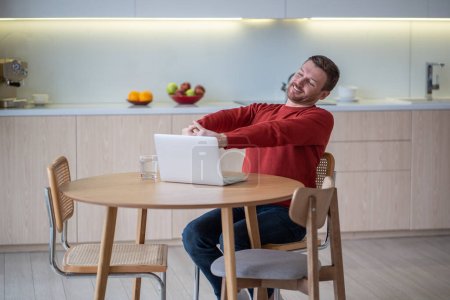 Téléchargez les photos : Homme indépendant positif prenant pause étirant les bras se reposant après avoir travaillé sur un ordinateur portable à la maison. gars agréable avec les yeux fermés souriant assis sur la cuisine avec ordinateur. Freelance, travail à distance, travail à distance - en image libre de droit