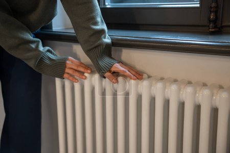 Photo for Woman warming hands near radiator at home after walking in cold winter weather, female touching barely warm battery during heating season, person near window checking heating system - Royalty Free Image