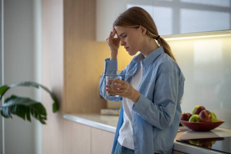 Photo for Woman feeling terrible pain in head drinking water on kitchen at home with closed eyes. Girl having headache, migraine, cephalalgia, premenstrual syndrome. Taking medications, painkillers concept. - Royalty Free Image