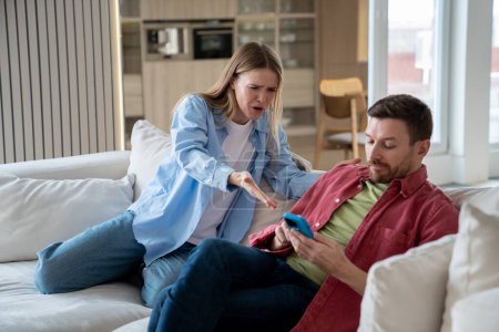 Photo for Angry irritated woman raising voice, shouting at demonstrative uncaring man sitting on sofa with mobile phone. Wife reproaches husbund in doing nothing, sitting and playing mobile games whole day - Royalty Free Image