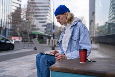 Photo for Trendy pensive hipster looking like freelancer, student, blogger, photographer, sitting in urban street with laptop, working, searching information, reading, texting, posting photos in social networks - Royalty Free Image