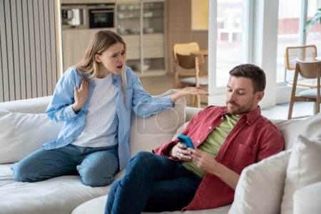 Photo for Indifferent ignoring man sits on sofa with mobile phone, playing games, scrolling social networks, reading news, keeping silence while offended irritated woman raises voice and shouts reproachfully - Royalty Free Image