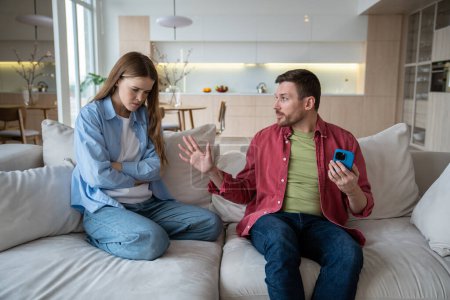 Photo for Angry annoyed woman listening to explanations of man holding smartphone, trying to hide messages, calls, information. Nervous wife suspects husband of unfaithfulness. Cheating, mistrust in family life - Royalty Free Image