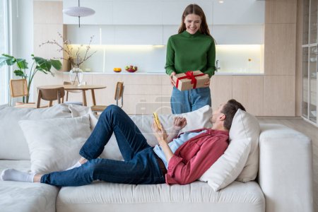 Photo for Loving woman presenting gift in cardboard box bound with ribbon to loved man lying on sofa. Male receiving present happy with unexpected surprise. Love, birthday. Tender relations, little family joys - Royalty Free Image