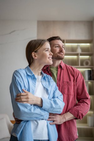 Photo for Romantic loving man and woman looking at window in contemplation, hugging with fondness, love and affection, smiling. Married couple of wife and husband dreaming of happy family life, future, children - Royalty Free Image