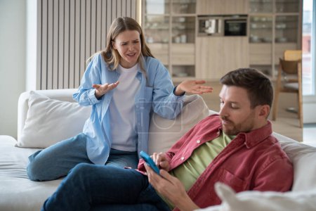 Photo for Hysterical excited woman blaming, shouting at uncaring indifferent lazy man sitting on sofa with smartphone. Wife regrets of wedding, creating family because husband doesnt satisfy expectations - Royalty Free Image