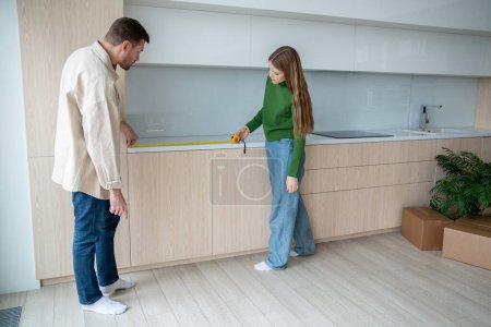 Téléchargez les photos : Homme concentré et femme concentrée mesurant la surface sur des armoires de cuisine à l'aide de ruban à mesurer. Couple familial déménageant dans un nouvel appartement, faisant des rénovations, modernisation du mobilier à la maison concept. - en image libre de droit
