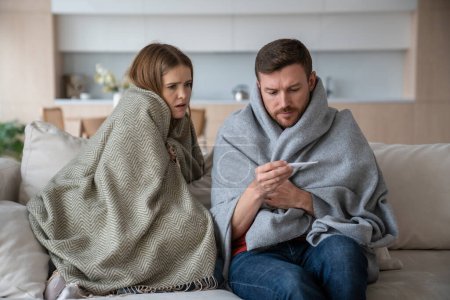 Photo for Family couple young man woman measuring temperature sitting on couch wrapping in plaids. Sad frustrated wife husband suffering from seasonal flu or cold. Worry about feeling unhealthy with influenza. - Royalty Free Image
