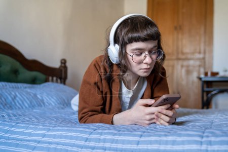 Téléchargez les photos : Amateurs de musique détendue adolescente écoute de la musique dans les écouteurs couchés dans le lit à la maison. Pensive adolescente écolière en utilisant un smartphone dans les écouteurs. Fan de musique profiter du temps libre passe-temps avec téléphone mobile - en image libre de droit
