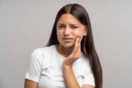 Photo for Unhappy teenage girl touching cheek with hand with painful face expression caused by toothache, dental pain, pulpitis, caries, periodontal inflammation. Teenager in need of stomatological treatment - Royalty Free Image