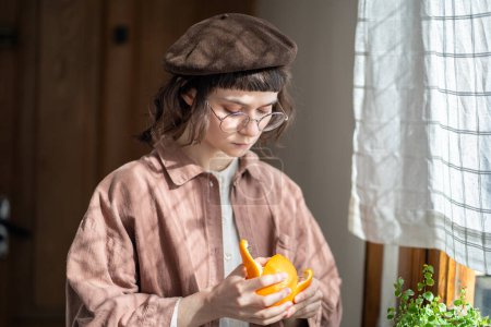 Photo for Pensive young woman in vintage style clothes, beret, eyeglasses peeling orange at home standing near window. Food snack, healthy eating nutrition concept. Useful product with vitamin C, sweet citrus. - Royalty Free Image