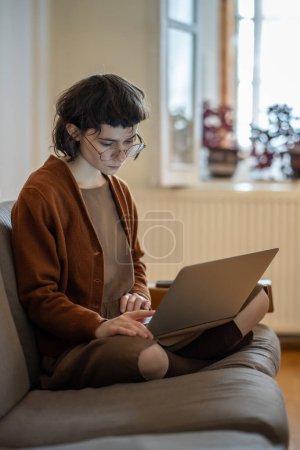 Téléchargez les photos : Fille indépendante concentrée assise à la maison avec un ordinateur portable sur les genoux, travaillant sur un nouveau projet sur ordinateur, pensant, faisant des devoirs sur un projet d'éducation à distance. Enseignement à domicile, travail indépendant, blogueur. - en image libre de droit