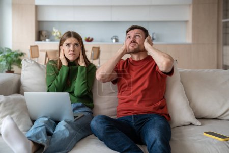 Photo for Annoyed unhappy couple sitting on sofa with vexed face expression, looking upward, closing ears because of loud sound, noise from neighboring flat. Emotional stress, discomfort caused by neighborhood - Royalty Free Image