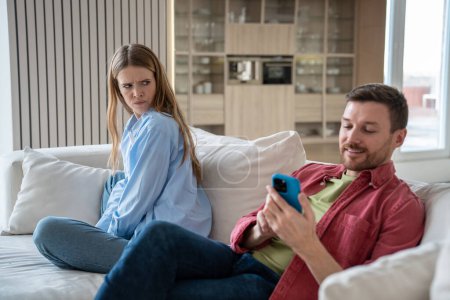 Wife angry at husband who looking at smartphone screen smiling sitting on couch at home. Family couple spouses having misunderstanding in relationships. Woman want to control private life of man.